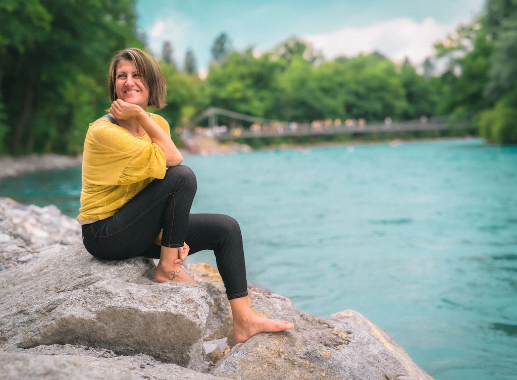 Maila Marchetta sitzt lächelnd an der türkisfarbenen Aare in Bern – ganzheitliche Begleitung für Frauen in der Lebensmitte